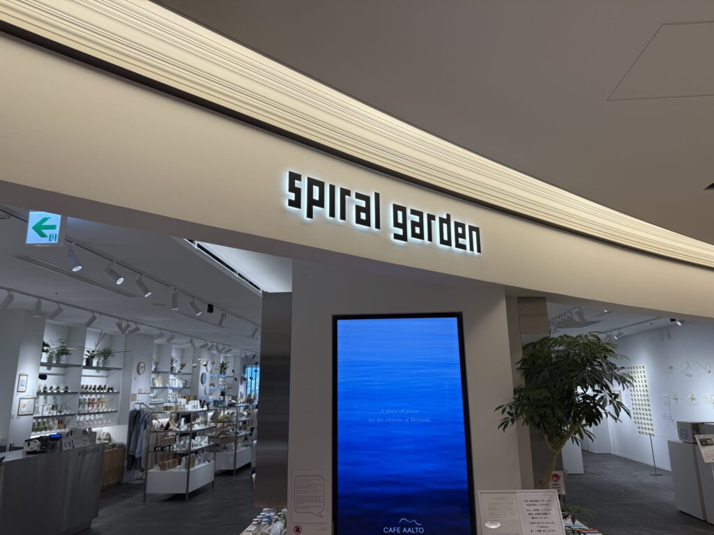 The backlit sign for 'SPIRAL GARDEN' above the entrance of a bright, white interior. Beyond the entrance is a lifestyle shop, and a blue digital sign indicates the presence of 'CAFE AALTO' within.