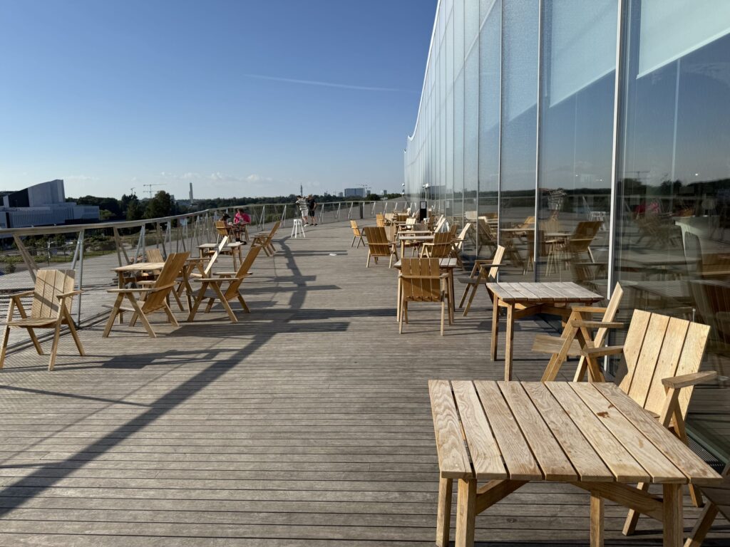 晴れた日の近代的な建物の広々としたウッドデッキのテラス。木製のテーブルと椅子が並び、ガラス張りの壁が空を映している。奥には数人の人々が見える。
