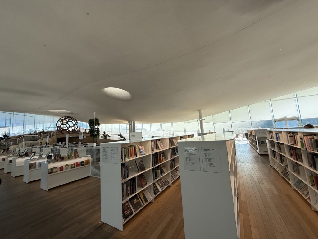 モダンな図書館の3階。白い波打つ天井の下に本棚が並び、大きな窓から光が差し込む中、人々が思い思いに過ごしている。