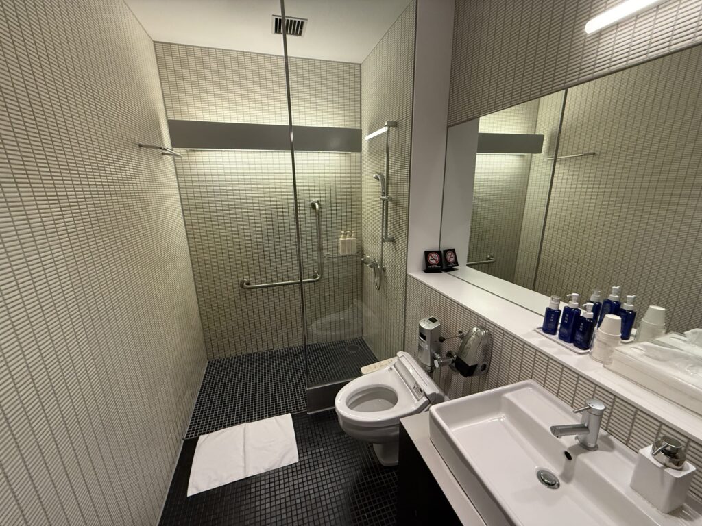 A modern shower room in an airport lounge. The compact space includes grey tiled walls, a glass-enclosed shower stall, a toilet, and a sink.
