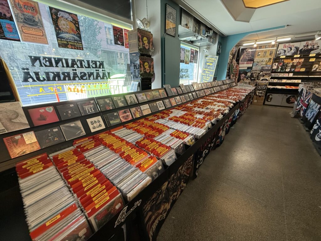 The interior of a record store, showing long black display bins packed with countless CDs. Posters adorn the walls and light streams in from the window.