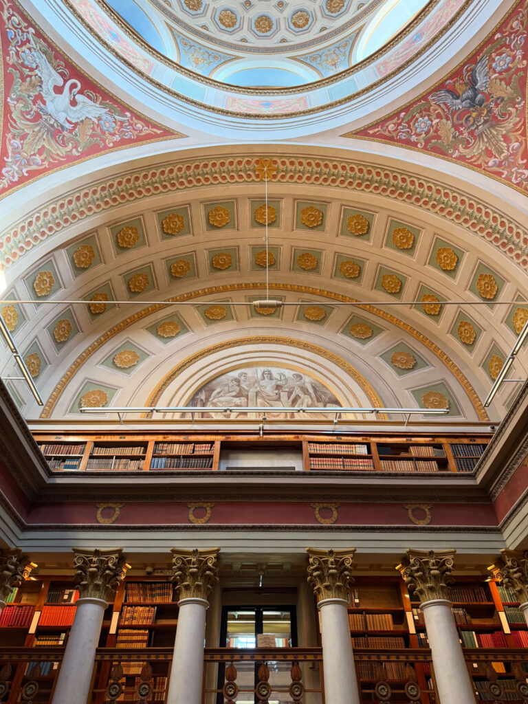 An upward view of the magnificent interior of the National Library of Finland, showcasing its ornate domed ceiling with frescoes, arches, and balconies lined with bookshelves.