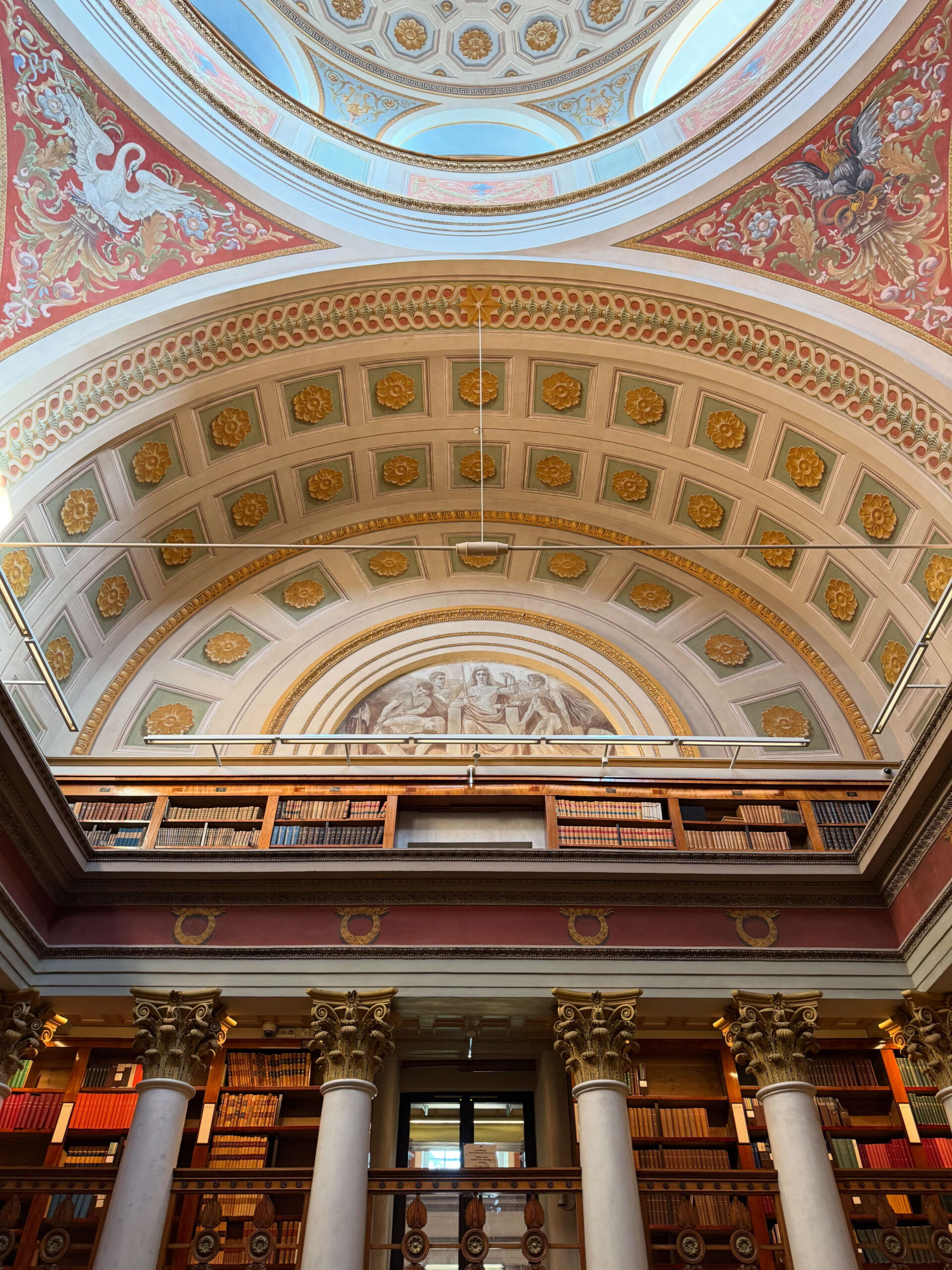 An upward view of the magnificent interior of the National Library of Finland, showcasing its ornate domed ceiling with frescoes, arches, and balconies lined with bookshelves.
