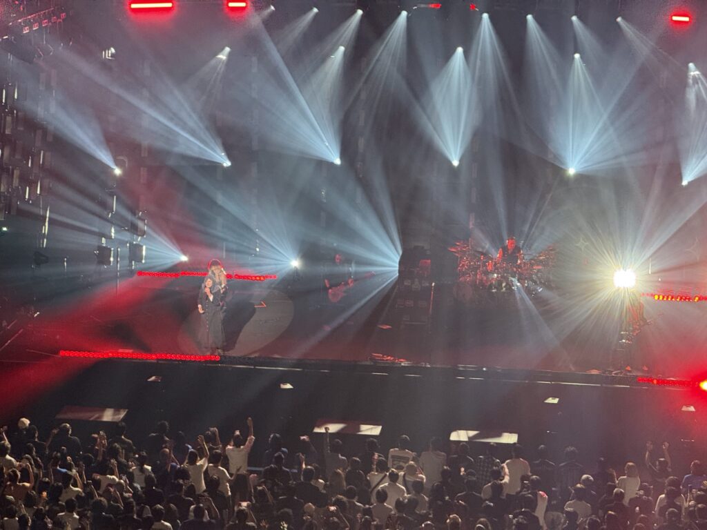 Billie Eilish and her band performing on a dark concert stage under powerful spotlights, with a large crowd in the foreground raising their hands in excitement.