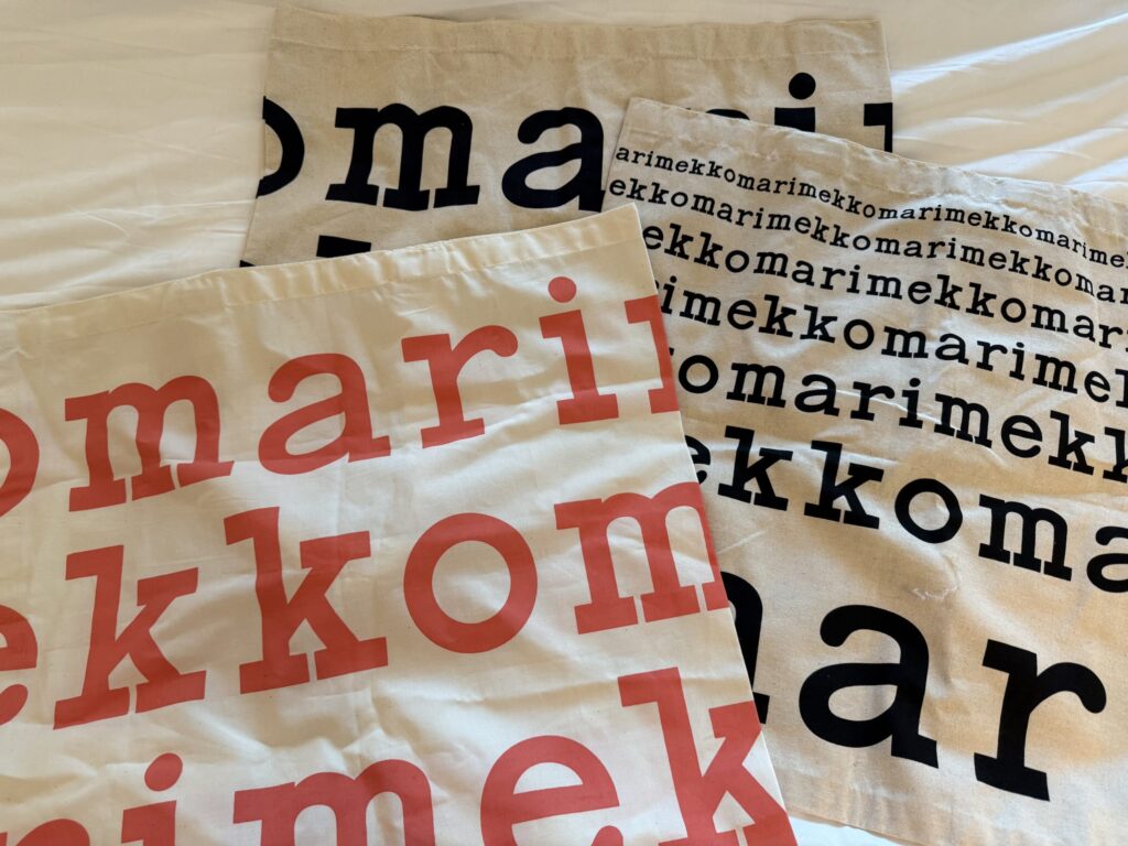 Three different Marimekko tote bags with various logo designs and colors, spread out on a white cloth.