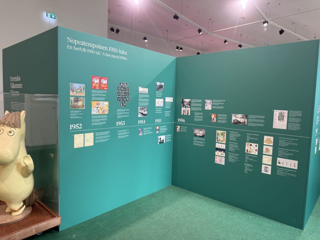 ムーミン展の内部。緑色の壁に1950年代のムーミンの歴史が年表で紹介されており、左側にはムーミンキャラクターの立体像が展示されている。