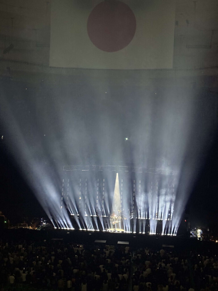 巨大なコンサート会場のステージ。天井には日本の国旗が大きく映し出され、ステージからは無数の白いスポットライトが客席に向かって放射状に伸びている。