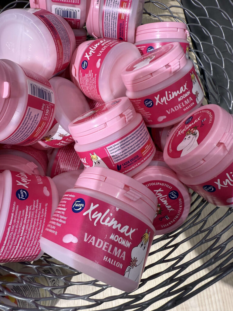 A wire basket filled with many pink containers of Fazer Xylimax Moomin xylitol pastilles, featuring illustrations of Moomin characters.