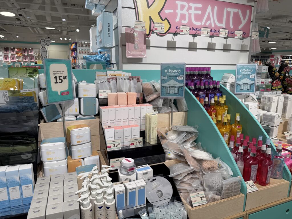 A view of a K-BEAUTY section in a store. A wide variety of Korean cosmetics and skincare products, such as face masks, serums, and creams, are displayed on turquoise shelves.