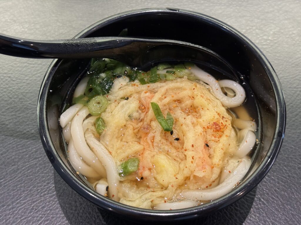 黒いお椀に入ったかき揚げうどん。ネギと七味唐辛子がトッピングされ、黒いレンゲが添えられている。