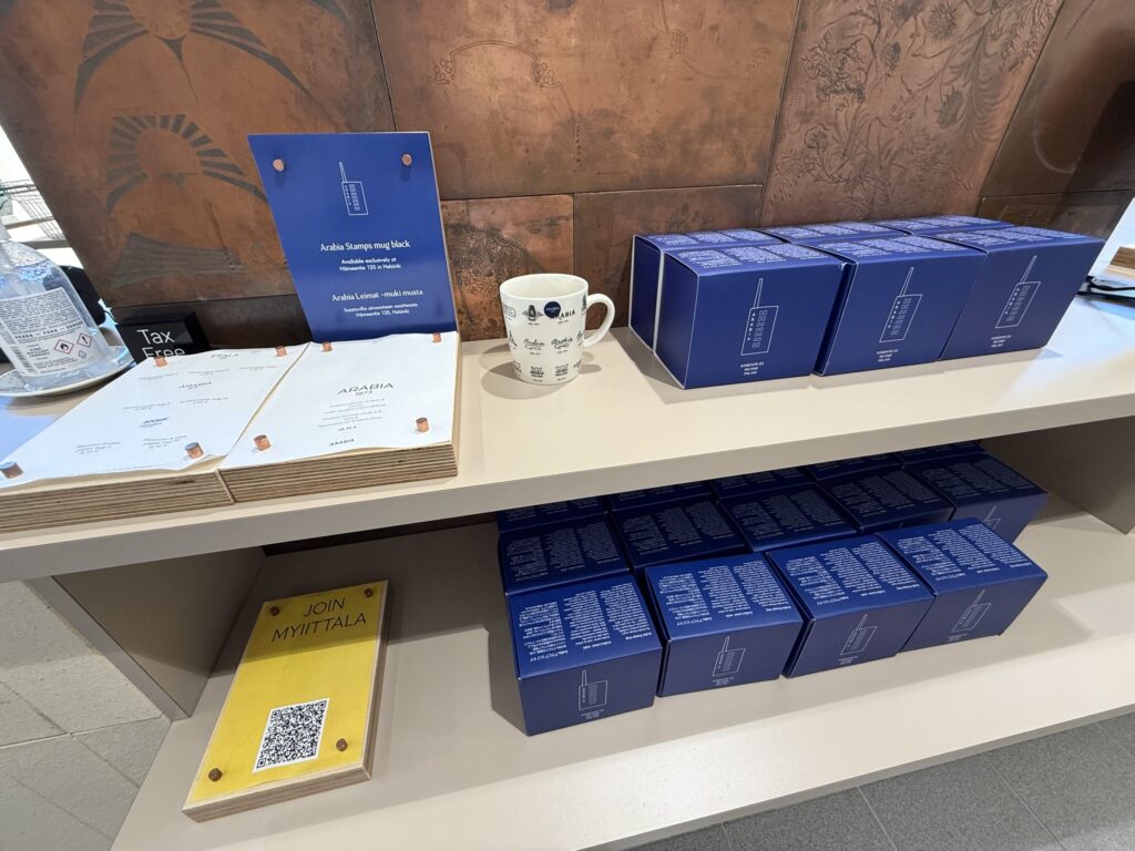 An in-store display at the Arabia Iittala Design Centre in Helsinki, showing the exclusive 'Arabia Stamps mug' and its blue packaging arranged on a shelf.