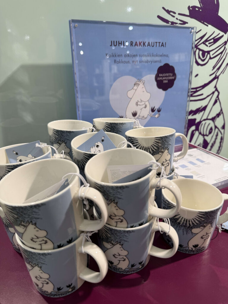 Light blue mugs with an illustration of Moomintroll and the Snorkmaiden hugging, stacked on a purple shelf.