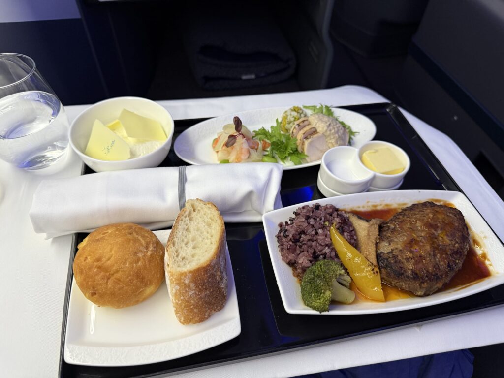 機内食のトレイに載せられた洋食。メインのハンバーグ、雑穀米、前菜、パン、バター、水が並んでいる。