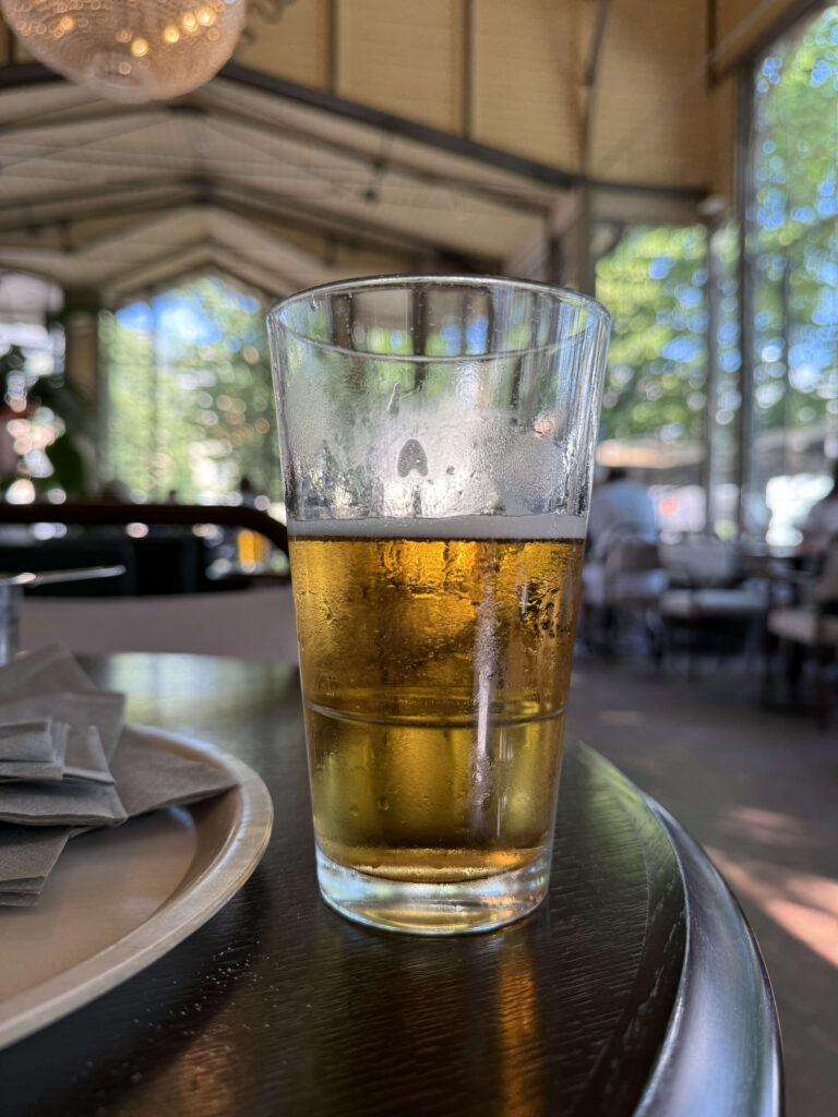 暗い木製のテーブルの上に置かれた、半分ほどビールが残ったグラス。背景には日差しが差し込む開放的なレストランがぼやけて写っている。