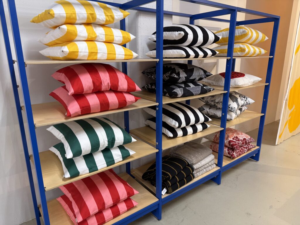 A blue shelving unit displays stacks of cushions with various striped patterns in different colors, including yellow and white, red and pink, green and white, and black and white.