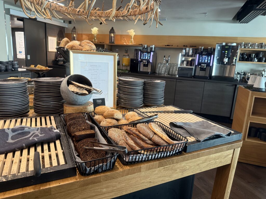 A breakfast buffet at a Lapland Hotel. A wooden counter displays various types of bread, including dark rye and rolls in baskets, next to stacks of dark plates. An antler chandelier hangs overhead.