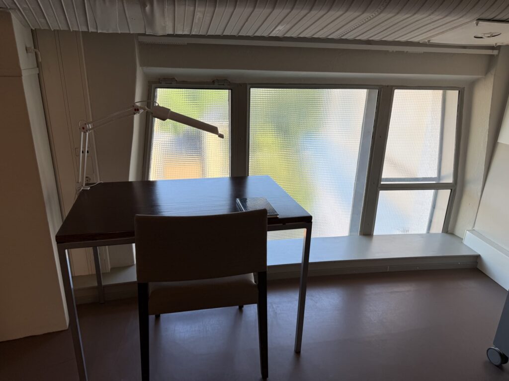 A quiet room with a desk and chair placed by a window. Soft natural light streams in through the patterned glass.
