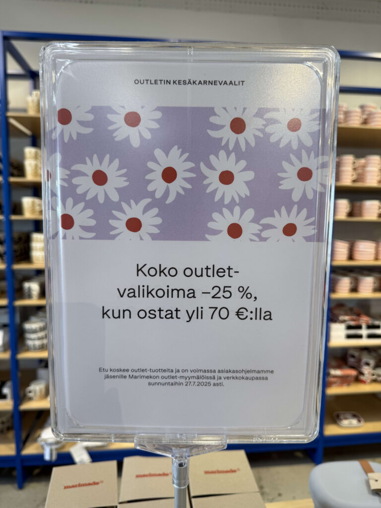 A sign for a promotional campaign inside a Marimekko outlet store. It features white daisies on a lavender background and text in Finnish announcing '25% off outlet items with a purchase of 70 euros or more.'