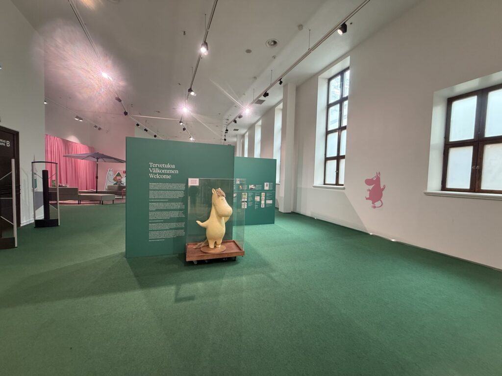 The Moomin exhibition hall. In a large space with green carpeting, there is a 3D statue of the Snorkmaiden in a glass case and a welcome board. A silhouette of Little My is painted on the wall.