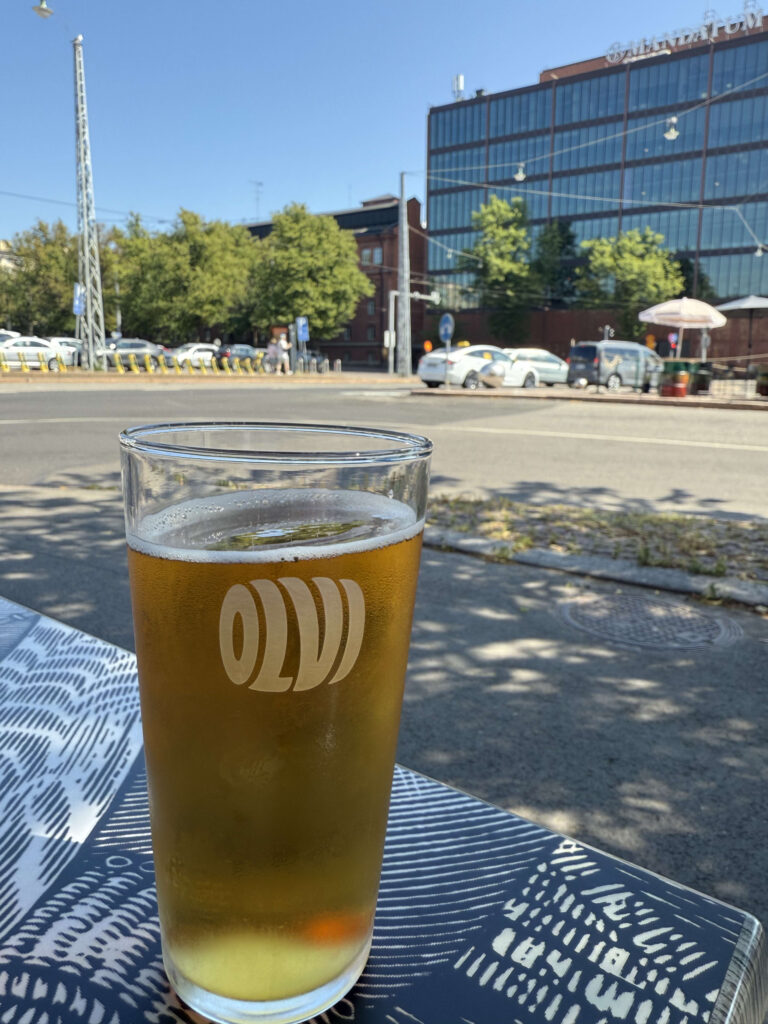 A glass of beer with the 'OLUT' logo on it, sitting on an outdoor table on a sunny day. In the background is a city street scene in Helsinki with cars, trees, and a modern building under a clear blue sky.