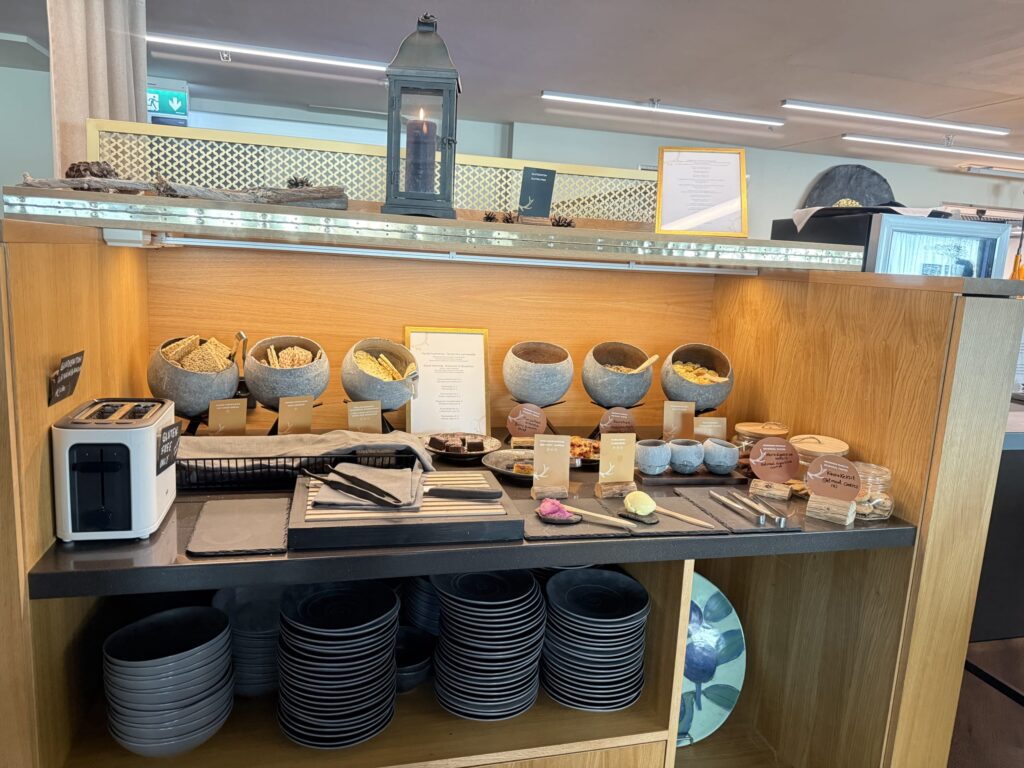 A dedicated vegan and gluten-free station at a hotel breakfast buffet, with a wooden counter holding a toaster, stone bowls of cereals and crackers, hummus, and stacks of black plates below.