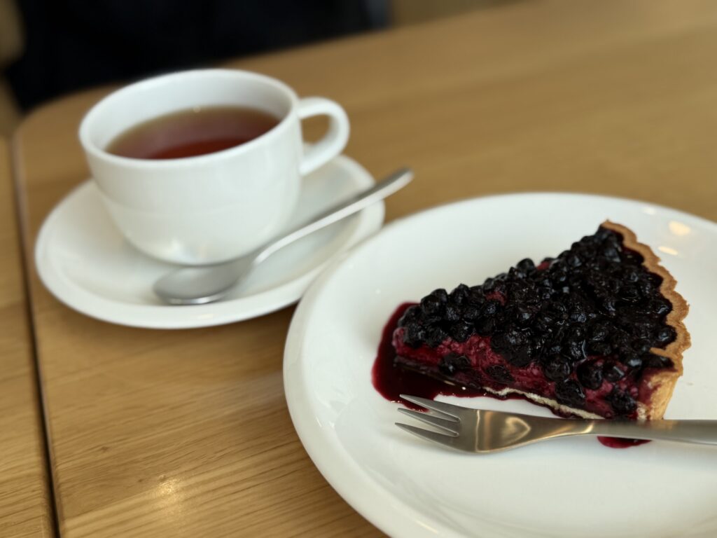 A slice of bilberry tart on a white plate with a fork, and a cup of tea in a white cup and saucer in the background, on a wooden table.