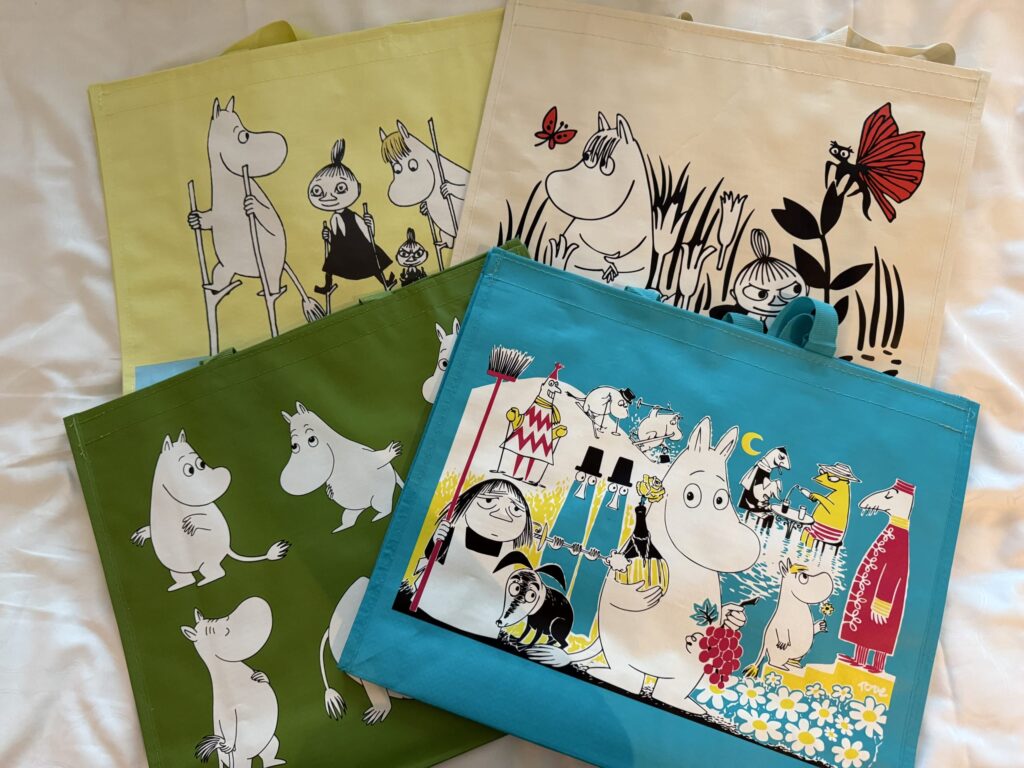 Four eco-tote bags in yellow, beige, green, and blue, featuring illustrations of Moomin characters, laid out on a white cloth.