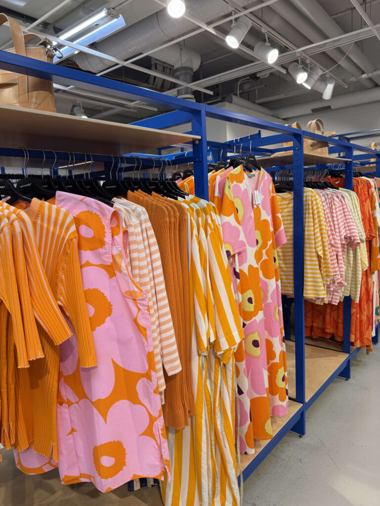 Inside a Marimekko store, a blue display rack is filled with colorful spring and summer clothes in shades of orange and pink, featuring iconic Unikko floral and striped patterns.