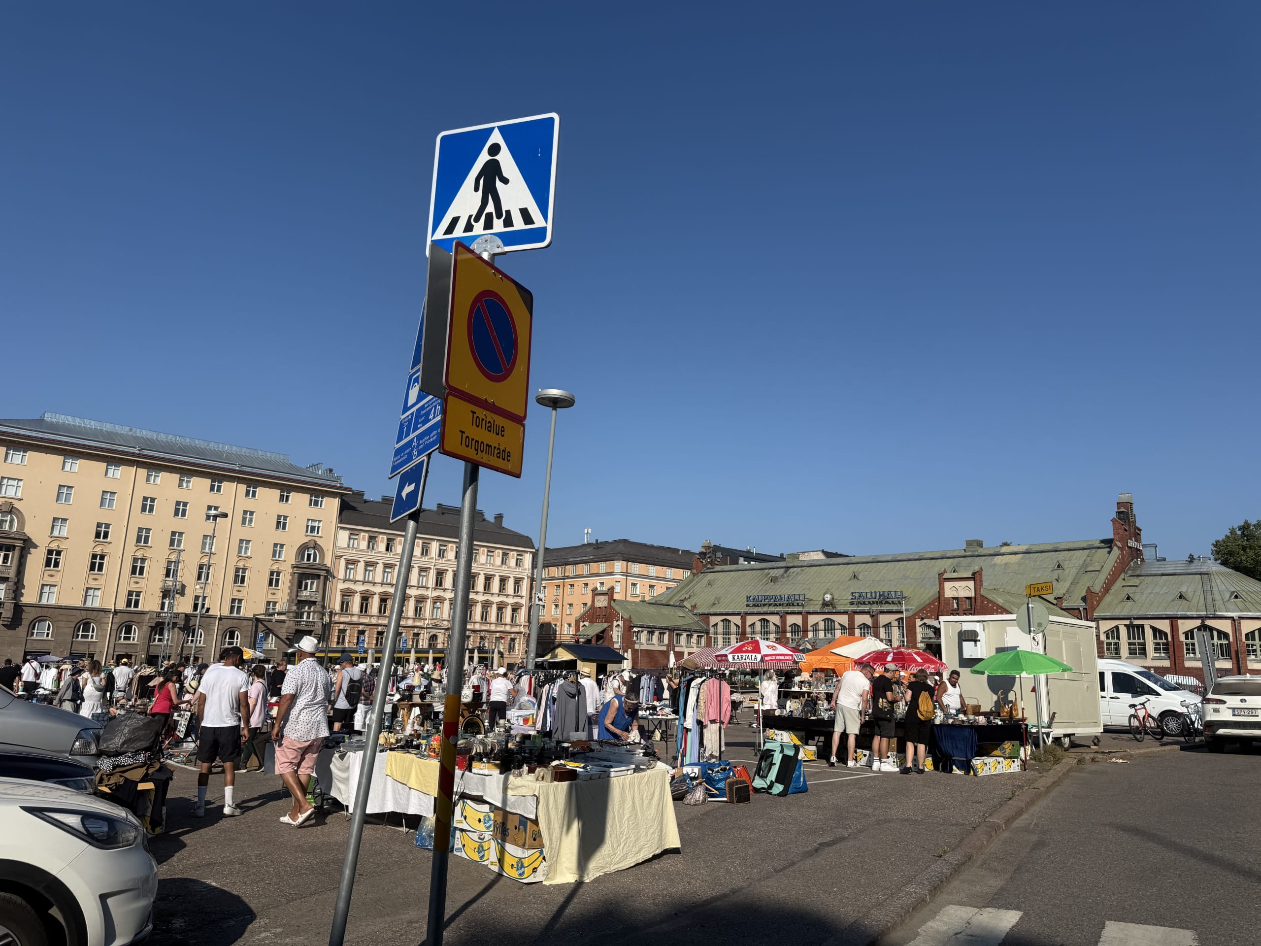 A bustling outdoor flea market at Hietalahti Square in Helsinki under a clear blue sky. Crowds of people browse stalls, with the historic market hall and apartment buildings in the background.