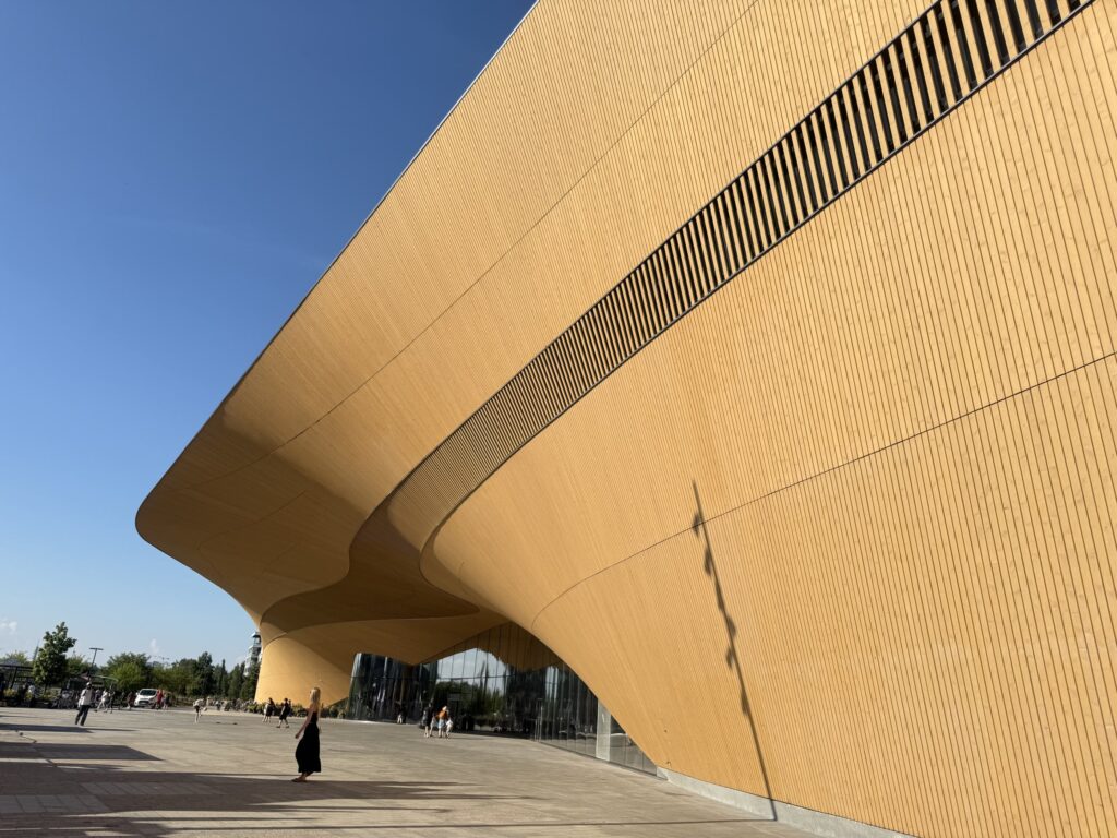 晴天の下、ヘルシンキ中央図書館Oodi（オーディ）の雄大な木製ファサード。下から見上げたアングルで撮影されており、建物のダイナミックな曲線と高さが際立っている。手前の広場には人々が行き交っている。