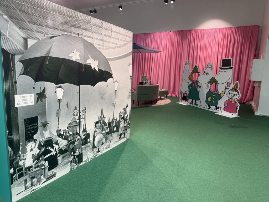 ムーミン展の会場風景。緑色の床の左手には大きな白黒写真のパネルが、右手奥にはピンクのカーテンを背景にムーミンキャラクターの顔はめパネルが設置されている。