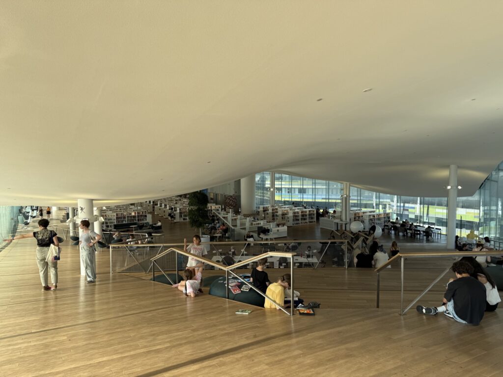 広々としたモダンな図書館の内部。波打つ白い天井と木の床が特徴的で、大きな窓から光が差し込む中、多くの人々が読書をしたり、床に座ってくつろいだりしている。