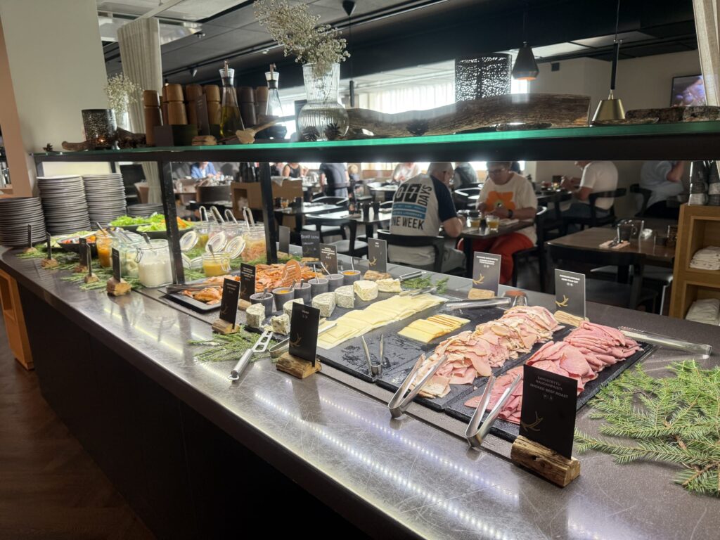 A wide variety of dishes including ham, cheese, smoked salmon, and salads displayed on a buffet counter in a restaurant, with people dining in the background.