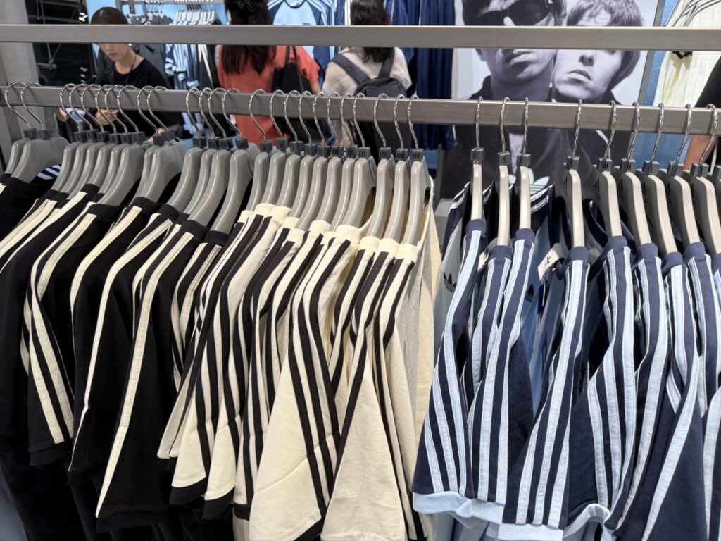 Adidas three-stripe t-shirts lined up on a rack in a clothing store. Shirts in black, off-white, and navy are hanging on display.