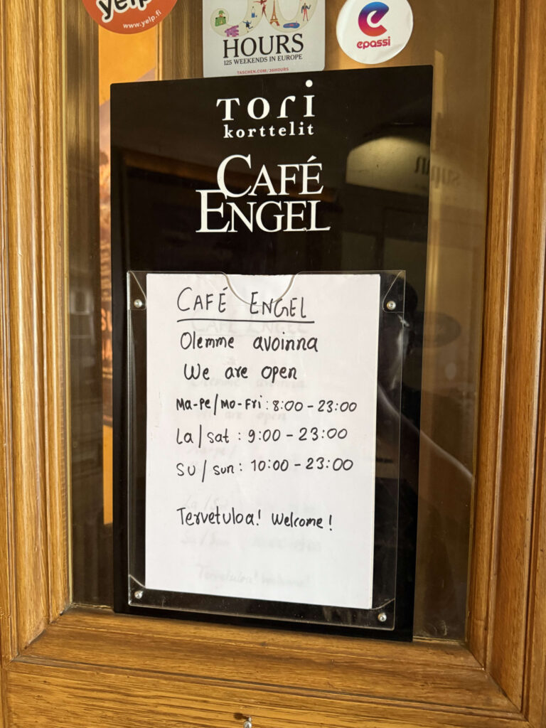 A handwritten sign showing the opening hours for Café Engel in Helsinki, posted on a wooden door.