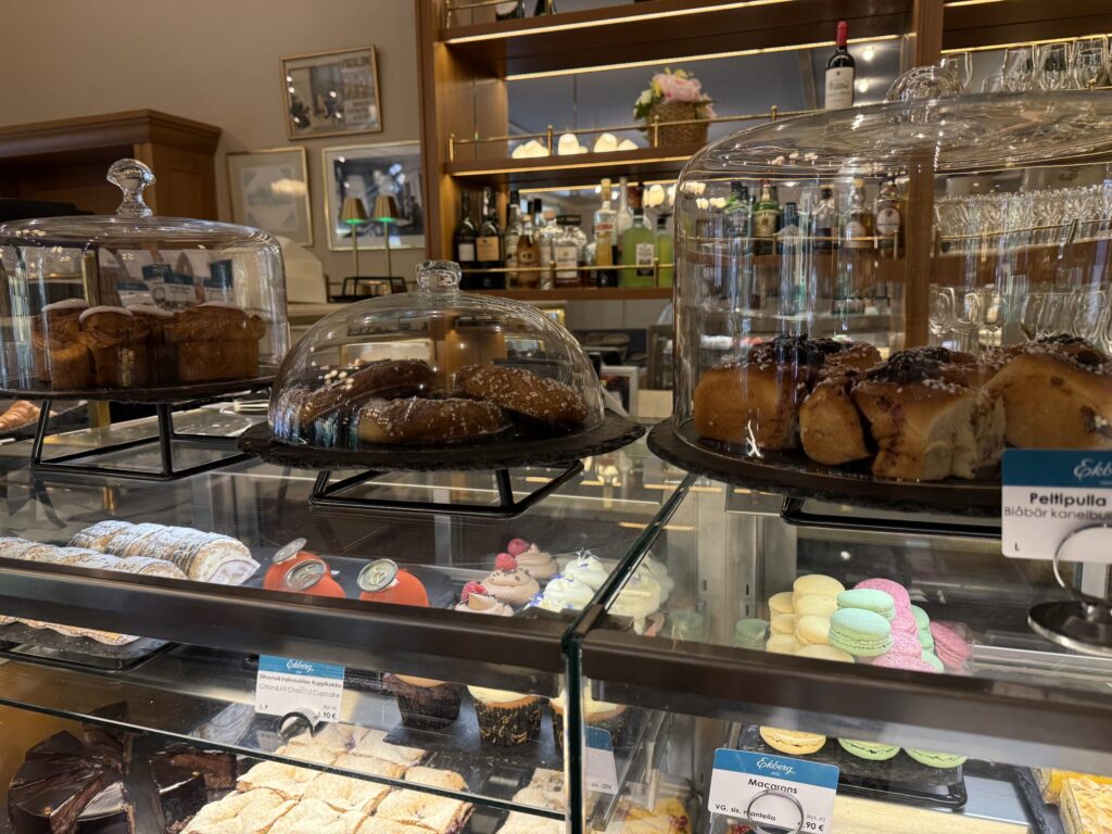 A glass display case at a cafe. The top shelf has cinnamon rolls and buns under glass domes, and the lower shelves are filled with colorful macarons, cupcakes, and chocolate cakes.