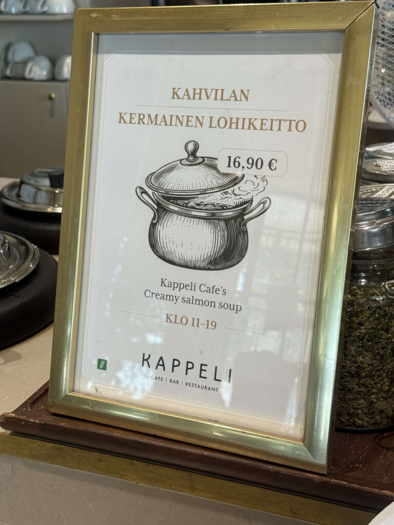 A menu sign in a gold frame at Kappeli Cafe. It advertises 'Creamy salmon soup' in Finnish and English, with an illustration of a soup pot and a price of €16.90.