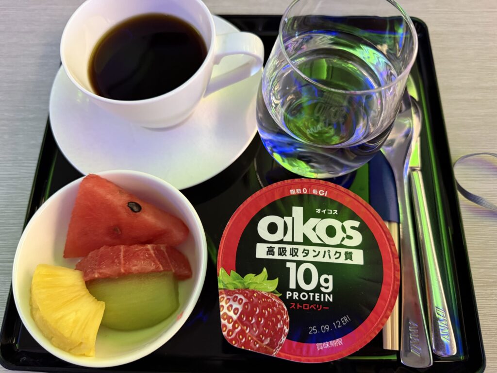 ANAの機内で提供された朝食。黒いお盆の上にコーヒー、水、カットフルーツ、ストロベリー味のオイコスヨーグルトが置かれている。