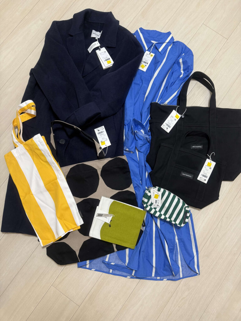 Several Marimekko items with tags still attached, including a navy coat, a blue and white striped shirt, black tote bags, and pouches, are laid out on a light wood-grain floor.