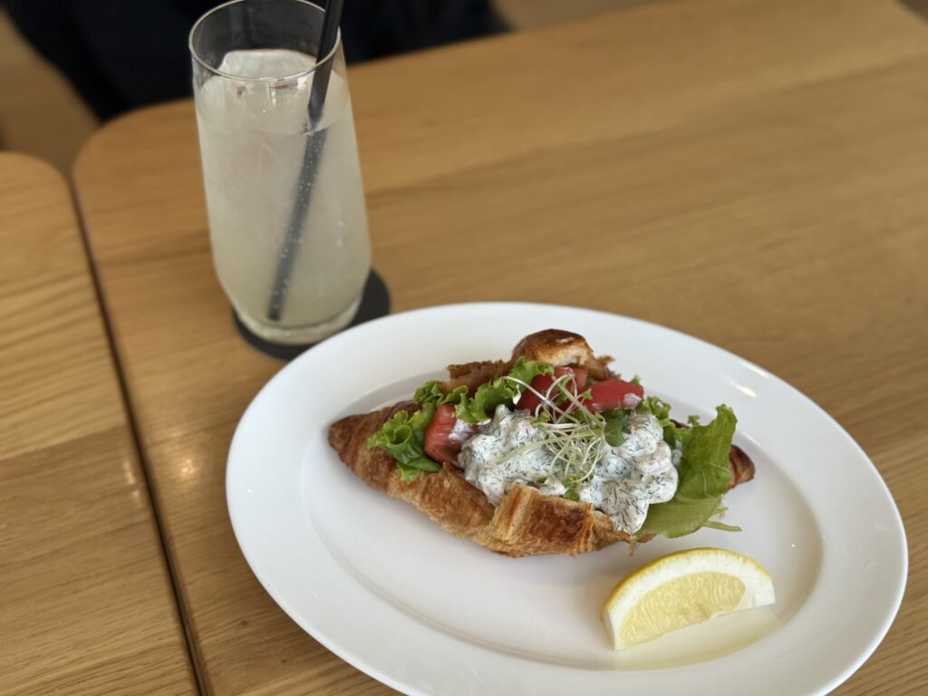 A shrimp croissant sandwich on a white plate and a glass of craft botanical squash on a wooden table.