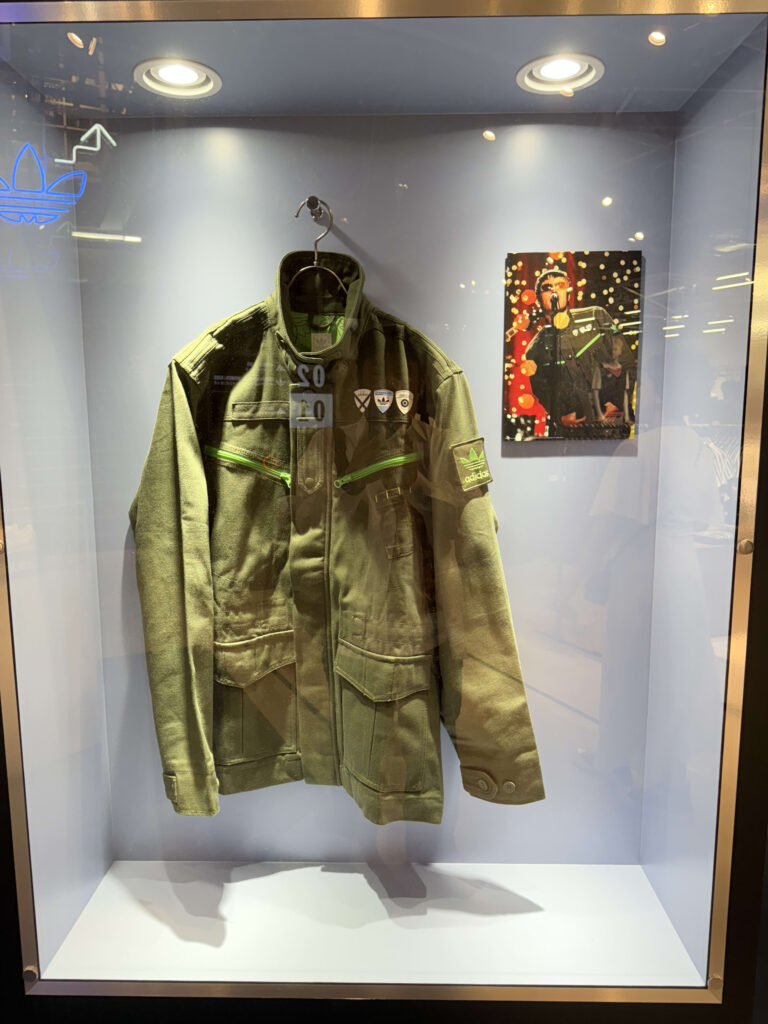 A green Adidas military-style jacket, worn by Liam Gallagher, displayed in a glass case. Next to it is a photo of him wearing the jacket during a live performance.