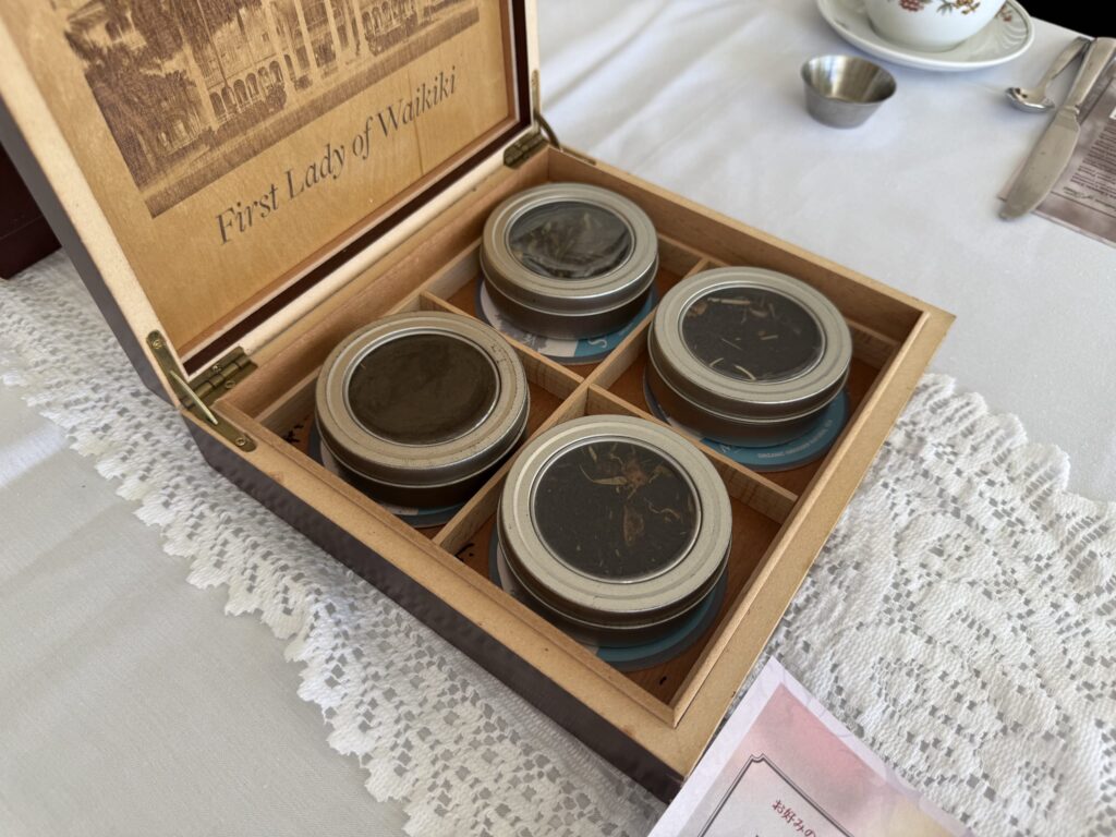 A wooden box containing four types of tea is placed on a lace tablecloth. The inside of the lid is engraved with 'First Lady of Waikiki'.