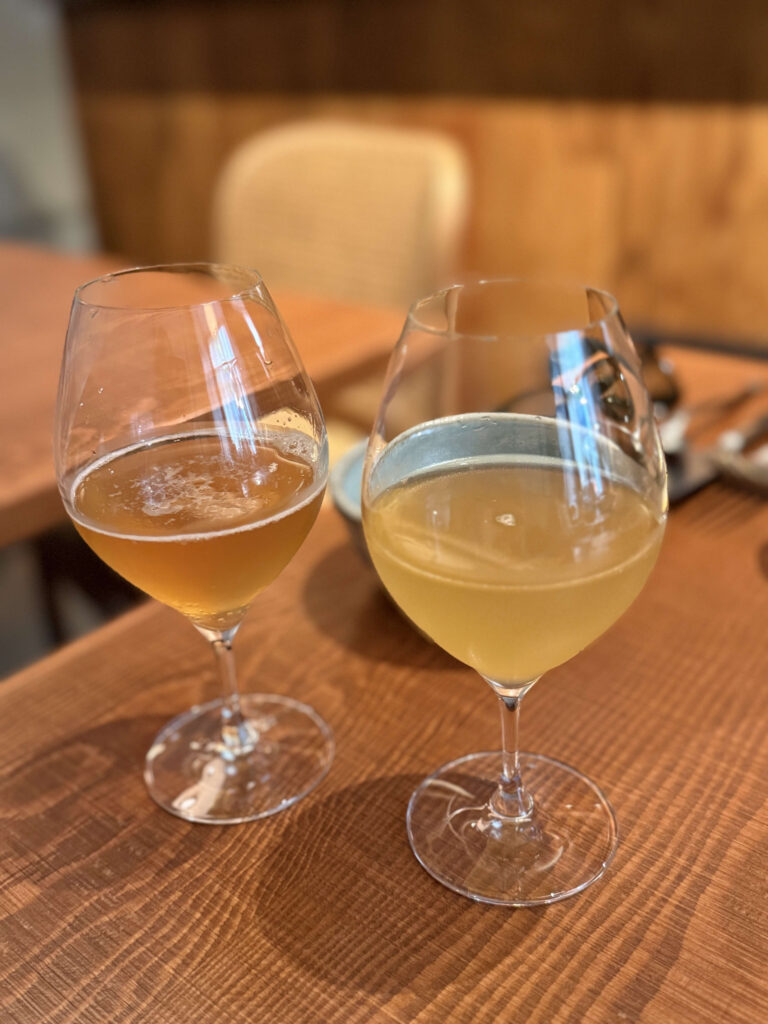 Two wine glasses on a wooden table. The left one is filled with amber-colored kombucha, and the right one with a pale yellow white tea sparkling drink.