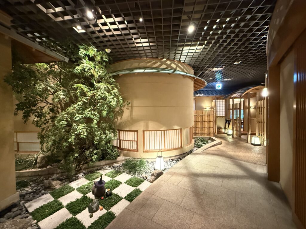 和モダンな内装の通路。左手には木が植えられた市松模様の坪庭があり、ハロウィンの飾りが置かれている。円筒形の壁と格子状の天井が特徴的。