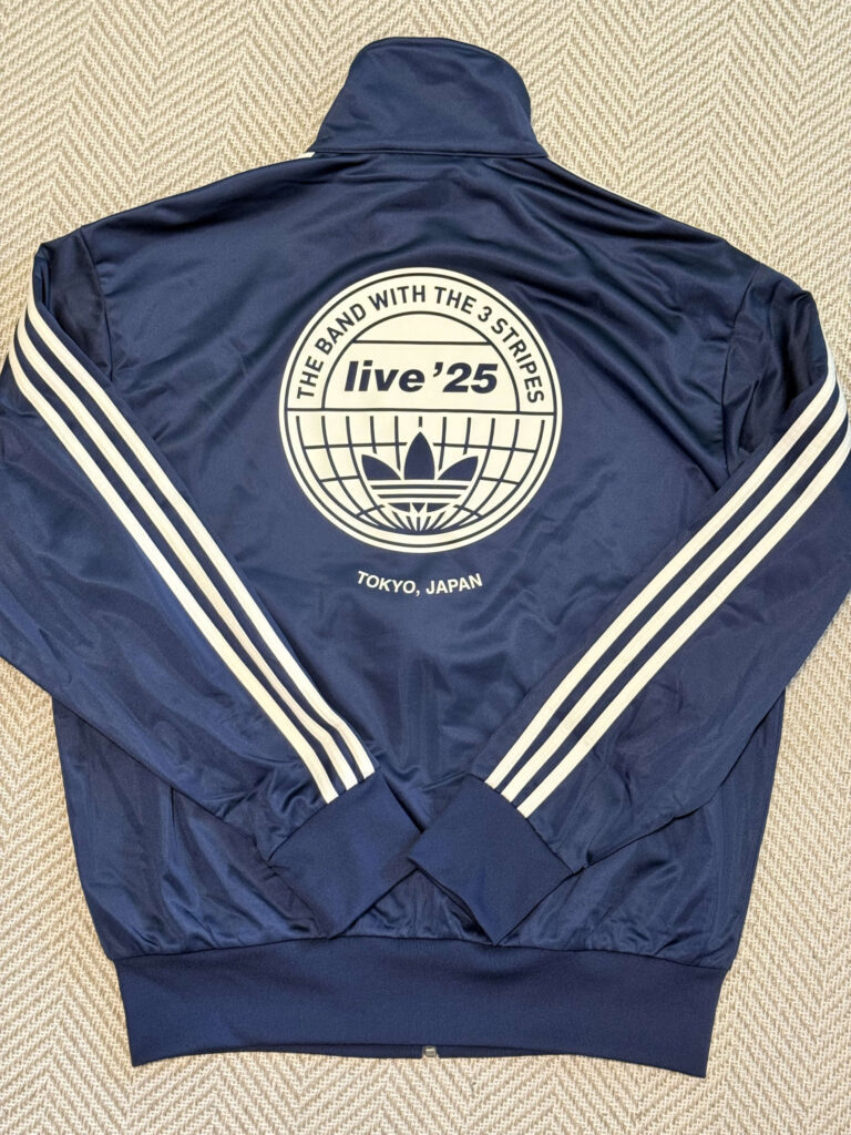 紺色のアディダス トラックジャケットの背面。中央に「live '25」と「TOKYO, JAPAN」の文字が入った大きな円形ロゴがプリントされている。袖には白の3本線が入っている。