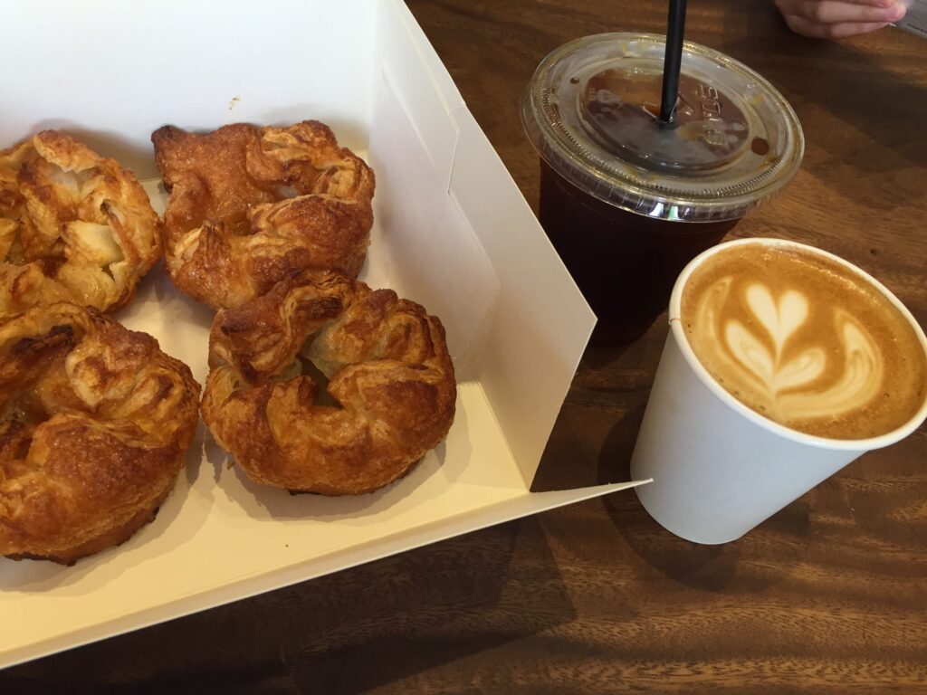 Four Kouign-amann pastries in a box, a caffè latte with heart latte art, and an iced coffee placed on a wooden table.
