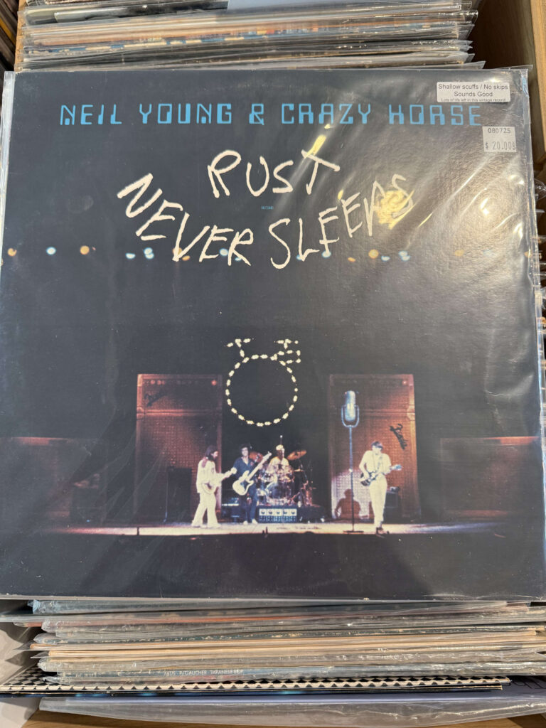 The vinyl record album 'Rust Never Sleeps' by Neil Young & Crazy Horse, stacked in a record crate. The album cover features a photograph of the band performing on stage.