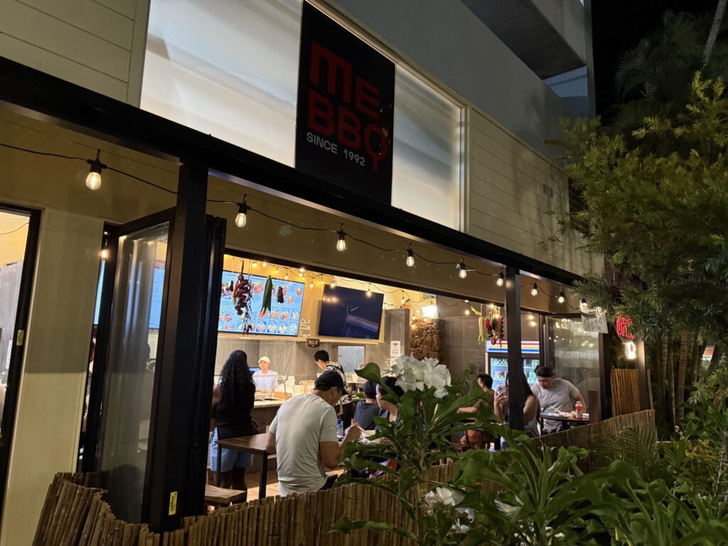 夜の「ME BBQ」レストランの外観。人々が食事を楽しむテラス席がストリングライトで温かく照らされている。