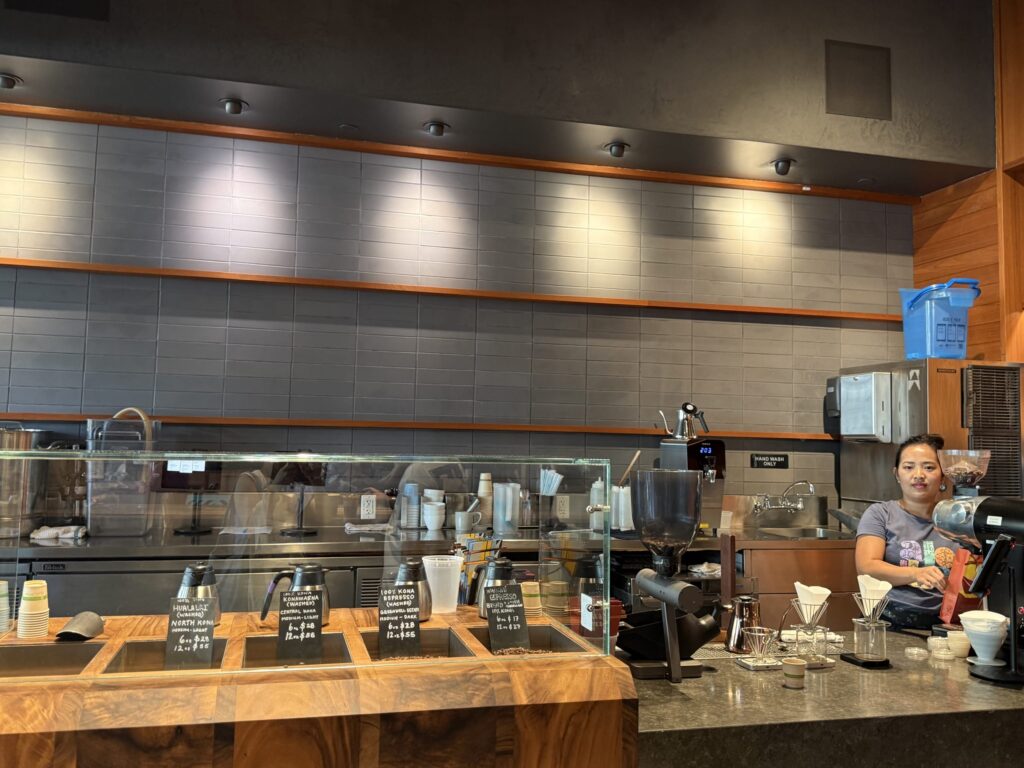 A female barista stands behind the counter of a modern coffee shop, where various types of Kona coffee beans are displayed for sale next to a pour-over coffee station.