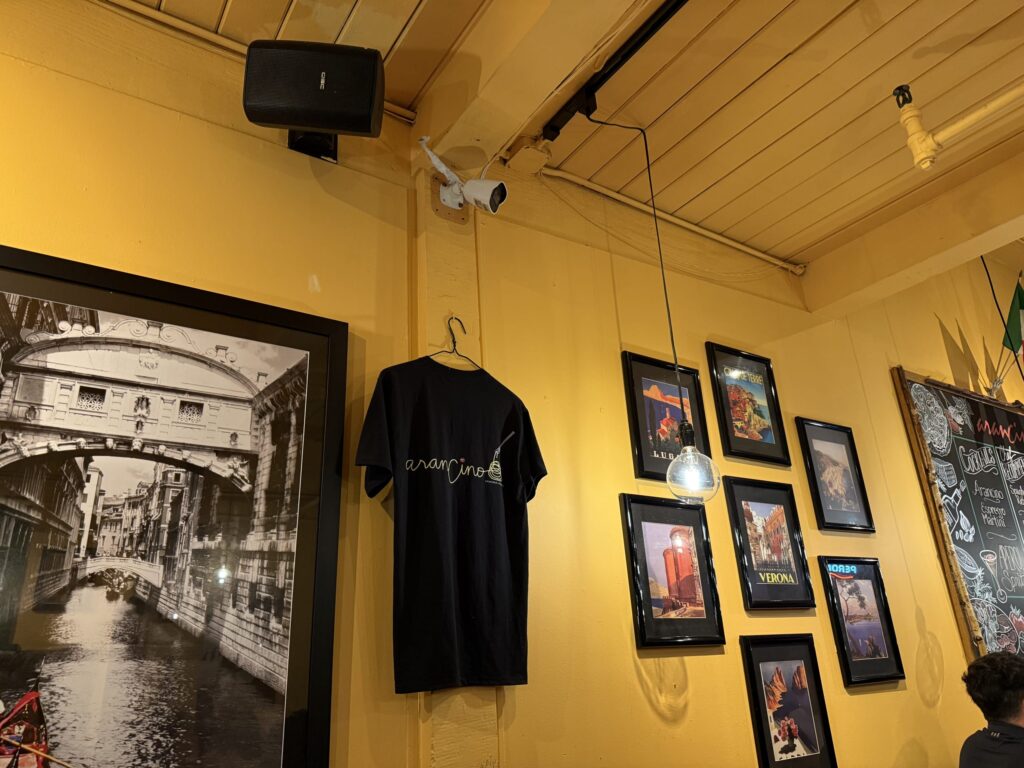 The interior of a restaurant with distinctive yellow walls and a wooden ceiling. The walls are decorated with a black-and-white photo of Venice, paintings of Italian landscapes, and the restaurant's logo T-shirt.